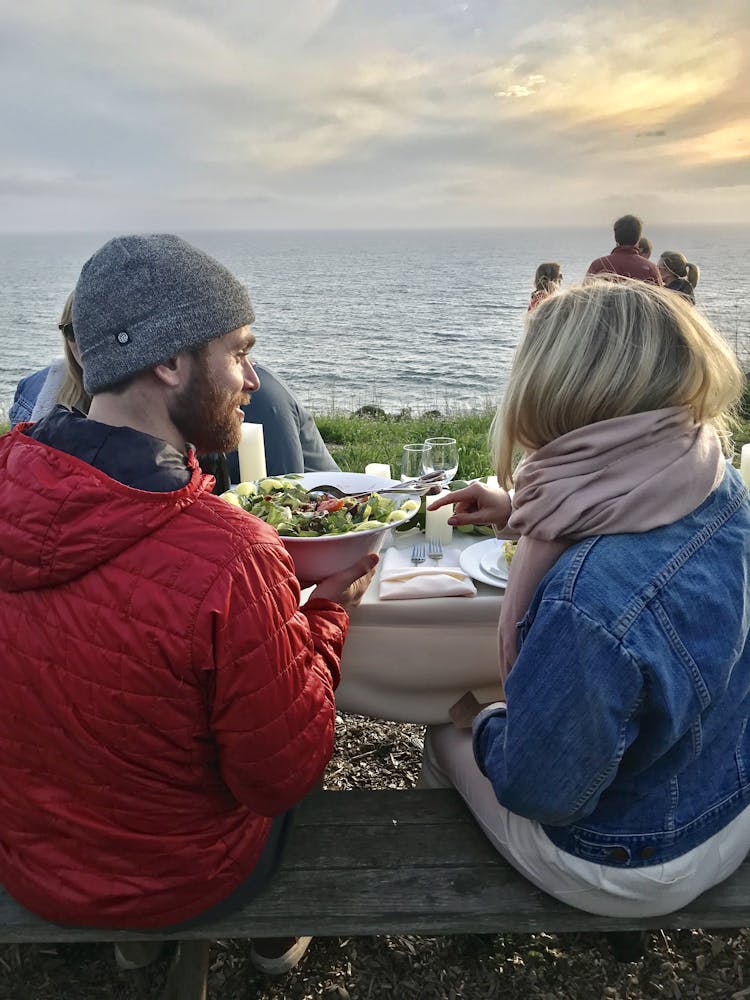 a man passing a woman a bowl of salad in front of a body of water